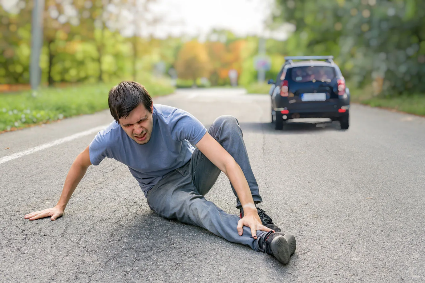 A man sitting on the road, holding his leg in pain, while a car drives away in the background. The scene implies a felony hit and run incident